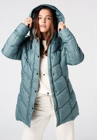 Gevoerde zeegroene jas met een verstelbare capuchon, voorzien van een diagonale quiltpatroon en zwarte drukknoopjes, gedragen over een lichtgekleurde trui.