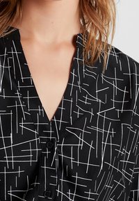 Blusa nera con scollatura a V, caratterizzata da un motivo geometrico bianco. Il tessuto è liscio, con chiusure a bottone lungo il davanti.