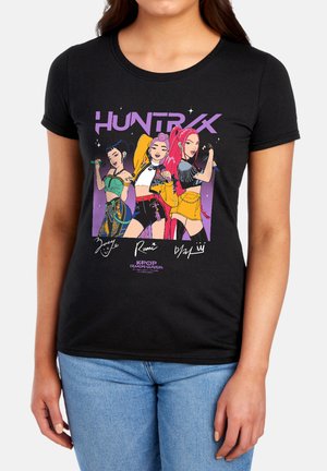 Three animated female characters in colorful outfits pose under "HUNTRIX" text on a black t-shirt worn with blue jeans.