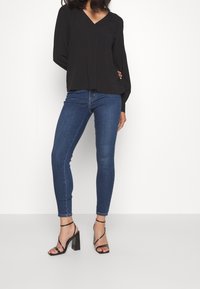 Svart långärmad blus med V-ringning, kombinerad med figurnära blå jeans och svarta sandaletter med klackar på en vit bakgrund.