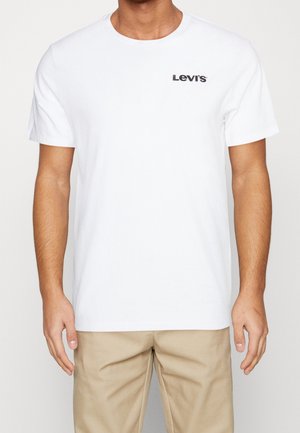 Mężczyzna w białej koszulce Levi's z logo po lewej stronie klatki piersiowej i beżowych spodniach, stojący z rękami swobodnie opuszczonymi wzdłuż ciała.