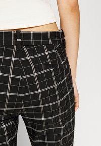 Pantalon à carreaux noir et blanc porté avec un haut blanc court, montrant une vue latérale arrière de la taille et des hanches.