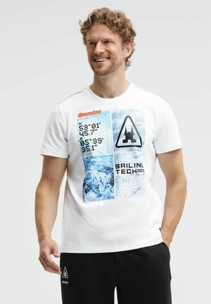 Man with curly hair smiles wearing a white t-shirt with nautical coordinates, ocean waves, and sailing-themed graphics.