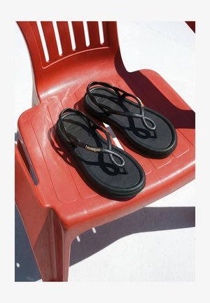 Schwarze Havaianas-Sandalen mit dünnen Riemen, die auf einem roten Plastikstuhl im grellen Sonnenlicht ruhen.