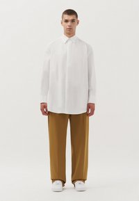 Witte blouse met lange mouwen en een puntige kraag, gecombineerd met losse mosterdkleurige broek en witte sneakers, minimalistisch ontwerp en een gladde textuur.