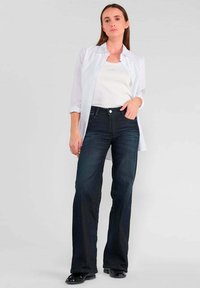 Un jean évasé bleu foncé avec une taille haute, un design à cinq poches et des dégradés subtils. Associé à un débardeur blanc ajusté et une chemise blanche ouverte.