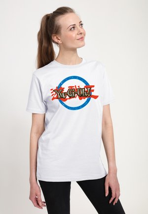 Ung kvinde med brunt hestehale iført hvid Yu-Gi-Oh! t-shirt og sorte bukser, kigger op og smiler let.
