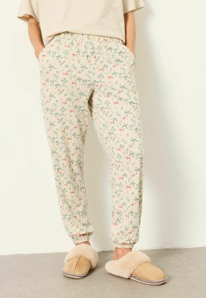 Persoon die lichtgekleurde pyjamabroek draagt met kleine dieren- en plantenmotieven, beige pantoffels met een pluizige voering, staand tegen een witte muur.