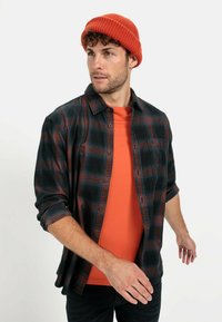 Sort og rød ternet skjorte over en orange t-shirt; model iført en orange strikket hue; afslappet pasform og knap-åbning.