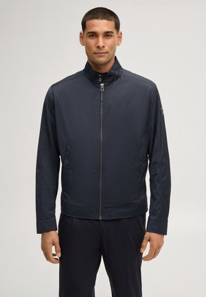 Mann trägt eine dunkelblaue Reißverschlussjacke mit Stehkragen und langen Ärmeln, steht vor einem schlichten hellgrauen Hintergrund.