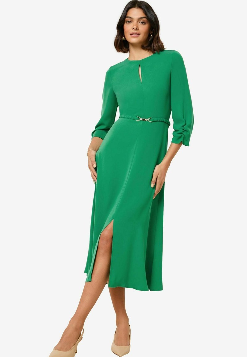 Abito midi verde con maniche lunghe, scollatura a chiave e uno spacco laterale. Presenta una vita con cintura e dettaglio della fibbia in tono oro.