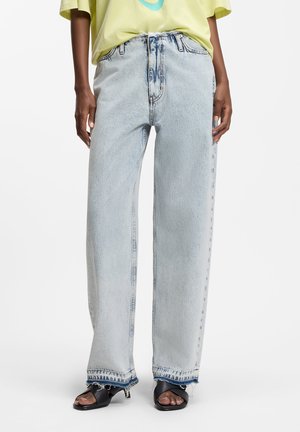 Άτομο που φοράει τζιν με ίσια μπατζάκια σε ανοιχτό μπλε χρώμα και ξεφτισμένα τελειώματα, κίτρινο πουκάμισο και μαύρα ψηλοτάκουνα σανδάλια ανοιχτών δαχτύλων, στέκεται μπροστά σε ένα λευκό φόντο.