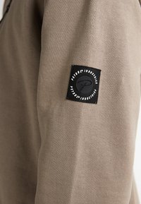Khaki mikina s měkkou texturou, která má na rukávu černou látkovou nášivku s oblíbeným kruhovým logem a textem.