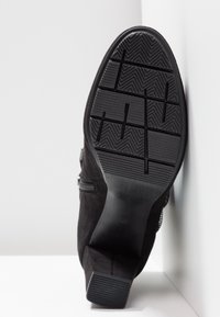 Semelle de botte à talon noir avec motif de grip texturé, inclinée contre une surface d'angle blanche montrant les détails de la bande de roulement et la forme du talon empilé.