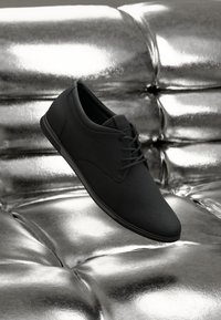 Zapato de vestir negro con cordones exhibido sobre una superficie de cuero plateado tufted brillante.