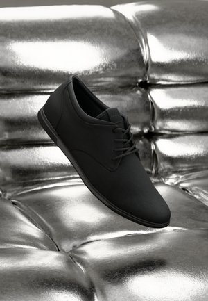 Zapato de vestir negro con cordones exhibido sobre una superficie de cuero plateado tufted brillante.