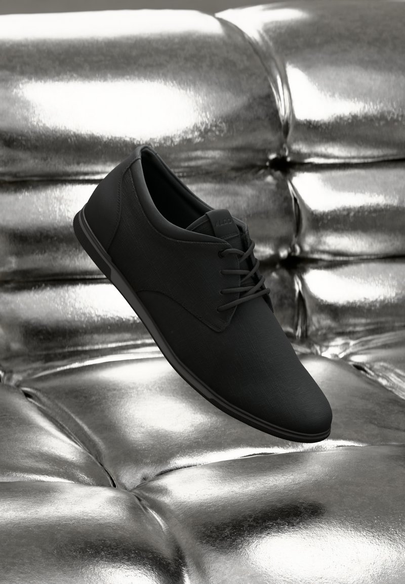 Zapato de vestir negro con cordones exhibido sobre una superficie de cuero plateado tufted brillante.
