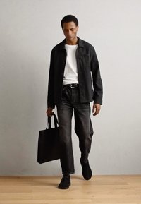 Veste noire sur t-shirt blanc, jeans foncés et chaussures noires. La personne porte un sac fourre-tout noir en marchant sur un sol en bois.