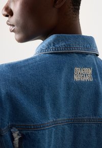 Giacca in denim dal taglio classico, con finitura blu, dettagli usurati e un logo ricamato in bianco sul colletto posteriore.