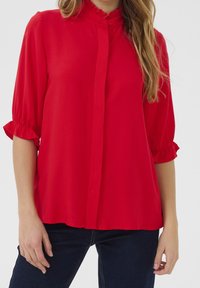 Blusa rossa con colletto arricciato, maniche a metà e chiusura a bottoni. Realizzata in tessuto leggero con una superficie liscia. Indossata con jeans scuri.