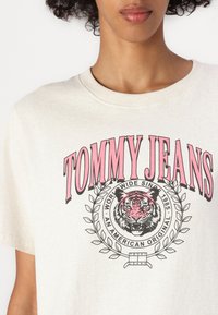Kremfarget t-skjorte med et grafisk motiv av et tigrehode, omkranset av et laurbærkransdesign, med rosa "TOMMY JEANS" tekst over.