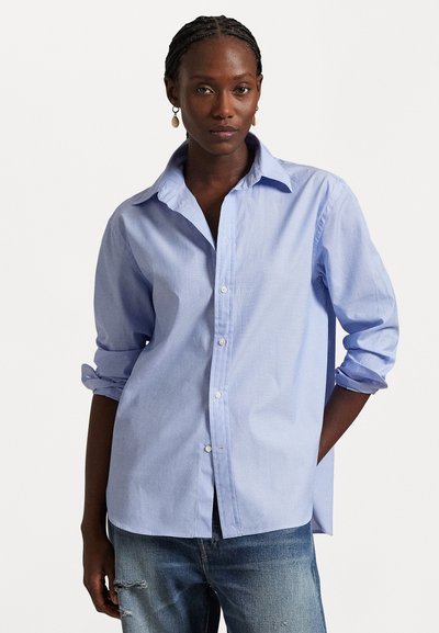 Femme aux cheveux tressés portant des boucles d'oreilles pendantes, une chemise bleu clair boutonnée avec les manches retroussées et un jean bleu déchiré, debout avec une main dans la poche.