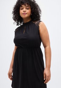 Μαύρο φόρεμα με δαντελένιες λεπτομέρειες στους ώμους, λαιμόκοψη με άνοιγμα τύπου keyhole και σούρα στη μέση, κατασκευασμένο από λείο, ελαστικό ύφασμα.