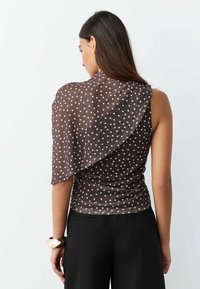 Brown polka dot top in sheer fabric, featuring a draped design and asymmetrical cut, paired with black pants and a statement cuff bracelet.