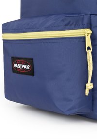 Zaino in nylon blu con una tasca con zip gialla e una patch nera con il logo Eastpak. Il cursore della cerniera presenta un dettaglio decorativo a nodo.