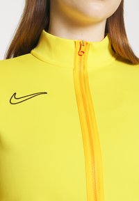 Keltainen vetoketjullinen takki, joka on valmistettu venyvästä materiaalista. Ominaisuuksiin kuuluu korkea kaulus ja oranssi vetoketju. Musta Nike-logo vasemmalla puolella rintakehää.