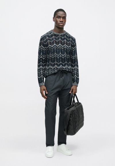 Juoda ir balta megzta megztinis su chevrono raštu, tamsiai pilkos siaurėjančios kelnės, balti sportiniai bateliai ir juoda dygsniuota rankinė.