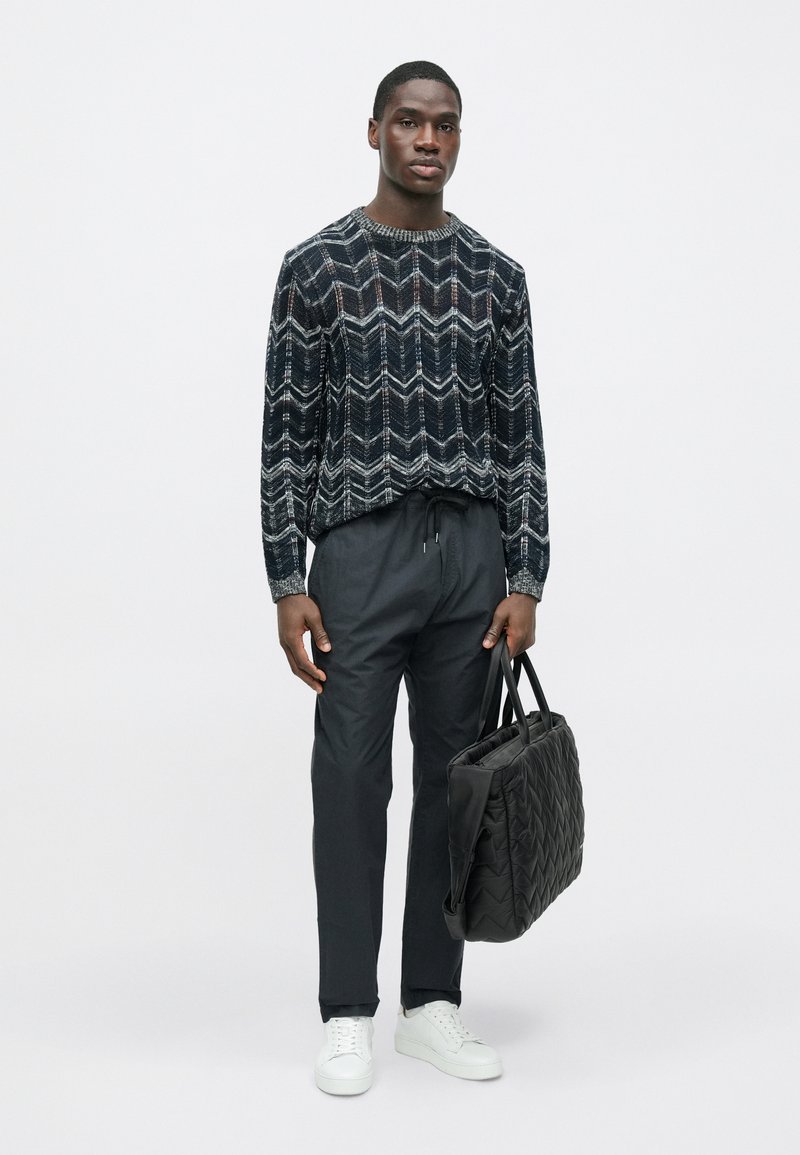 Pull en maille à motif chevron noir et blanc, pantalon fuselé gris foncé, baskets blanches et sac cabas matelassé noir.