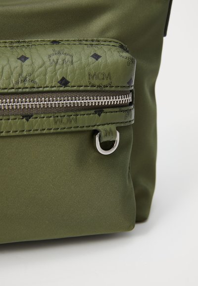 Geantă de culoare verde măslinie cu un accent din piele texturată, prezentând un model în diamant; include un fermoar argintiu și un detaliu hardware circular.