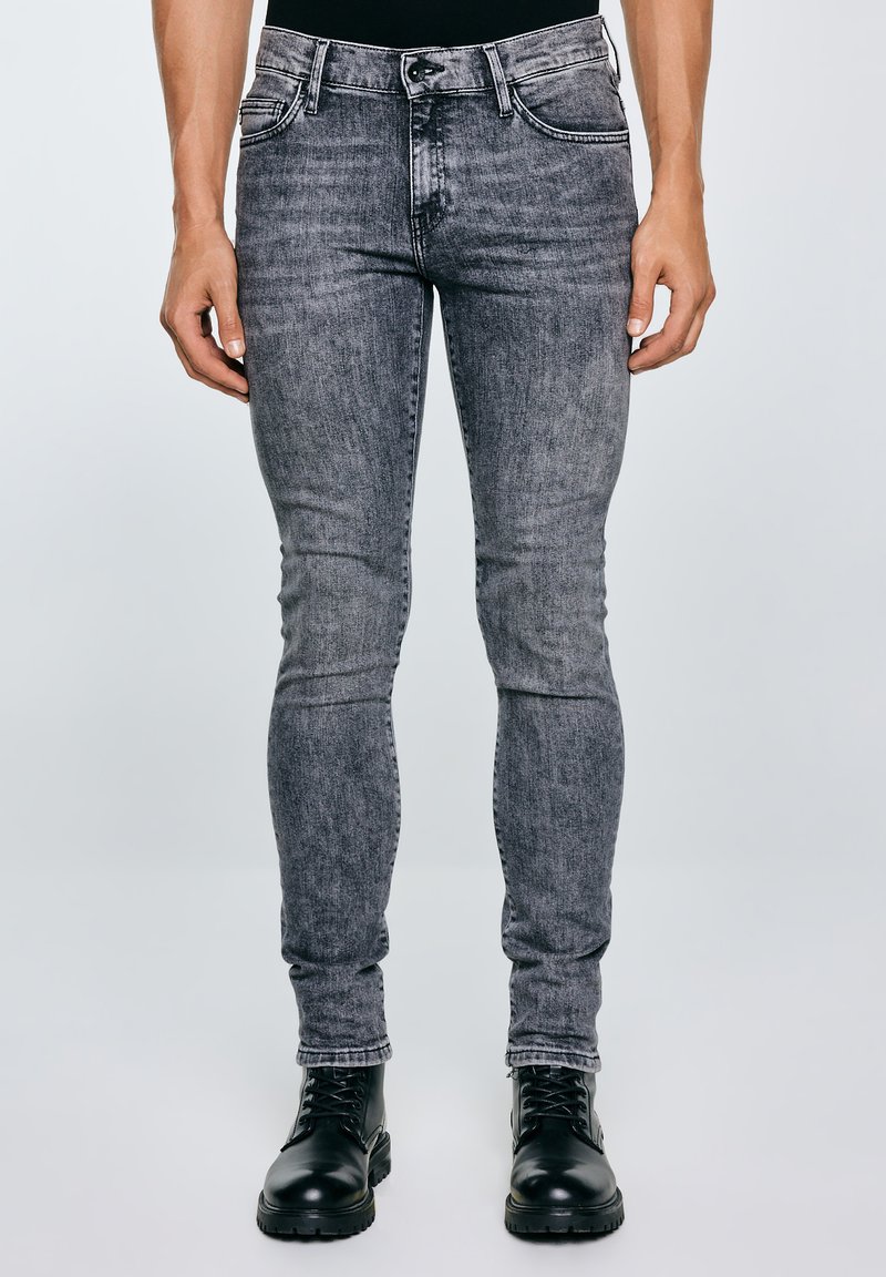 Õhukese lõikega tuhmhallid teksad, mida kantakse koos mustade pits-up kummikute ja seisva inimesega ühevärvilise tausta ees.