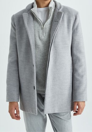 Uomo con soprabito grigio chiaro sopra maglione a mezza zip a coste e T-shirt bianca, con pantaloni grigio chiaro abbinati, in piedi contro uno sfondo neutro.