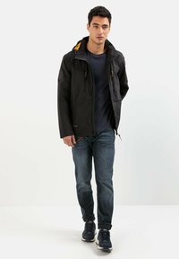 Sort vandtæt jakke med orange inderside, frontlynlås og lommer, iført over en mørk skjorte og blå jeans, suppleret med mørke sneakers.