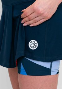 Skort azul marino con un diseño en capas, que presenta un parche de logo blanco. Debajo tiene pantalones cortos con patrones en tonos de azul y negro, mostrando formas geométricas.