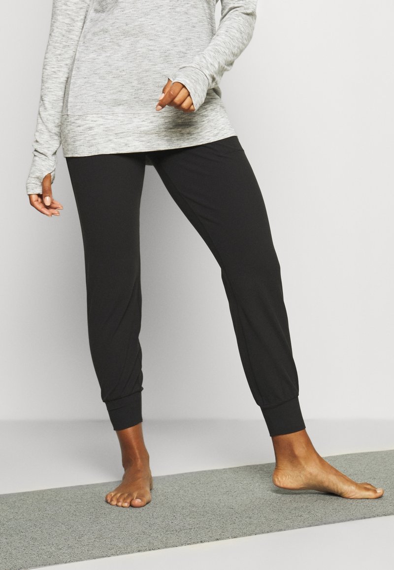 Personne debout pieds nus sur un tapis de yoga gris, portant un pantalon de jogging noir et un haut à manches longues gris clair, sur un fond uni.