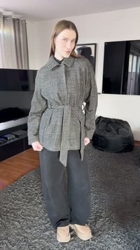 Dėvima didelio dydžio languota pilka ir juoda marškiniai su diržu ties juosmeniu, ilgomis rankovėmis ir smailiomis apykaklėmis, derinami su laisvomis juodomis kelnėmis ir smėlio spalvos batais.