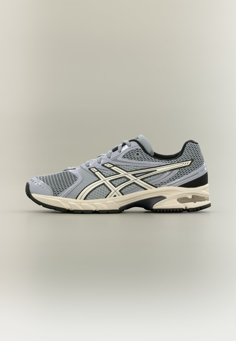 Grauer Sportschuh mit Mesh-Obermaterial, weißen Akzenten, schwarzer Ferse und profilierter Sohle. Verfügt über ein Schnürsystem und ein atmungsaktives Design.