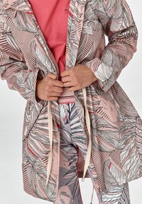 Lekki różowy płaszcz przeciwdeszczowy z czarnymi wzorami liści, ze sznurkiem w talii i dwoma przednimi kieszeniami, nałożony na różową koszulkę i wzorzyste legginsy.