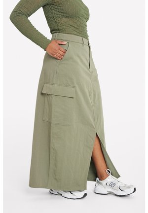 Woman wearing olive green cargo maxi skirt with side slit and white New Balance sneakers, hand resting in pocket.