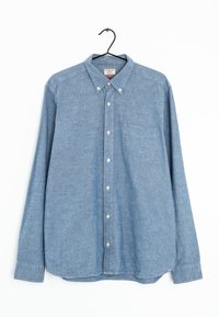 Chemise bleu clair à manches courtes en tissu chambray, avec une poche poitrine, un col pointu et des poignets ajustables. Suspendue à un crochet noir.