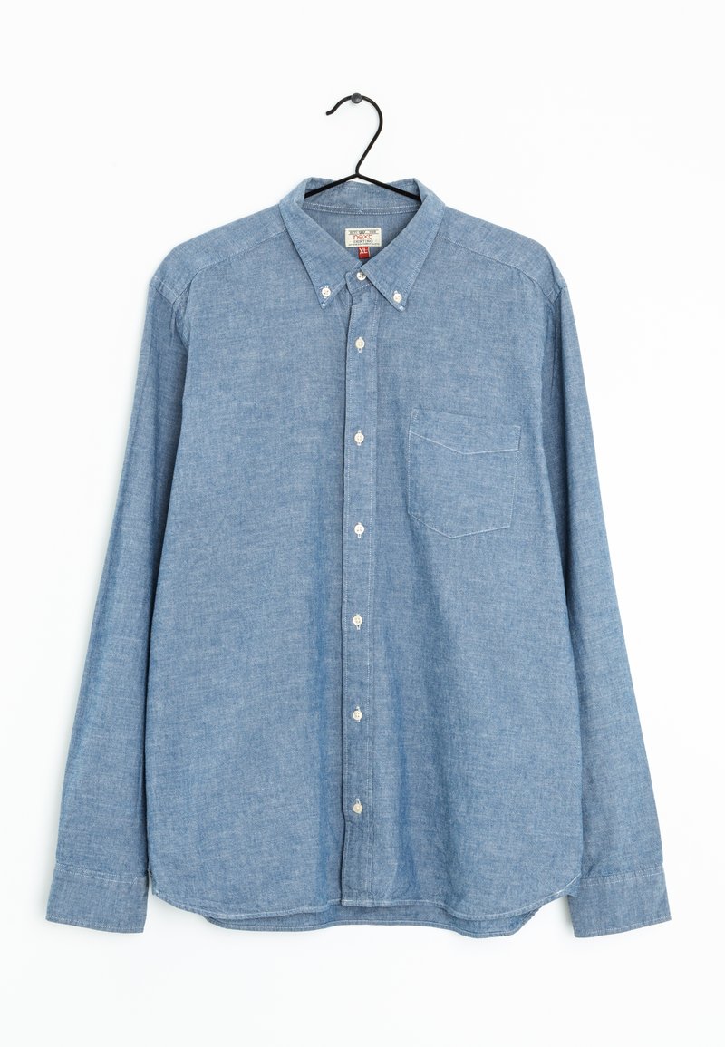 Chemise bleu clair à manches courtes en tissu chambray, avec une poche poitrine, un col pointu et des poignets ajustables. Suspendue à un crochet noir.
