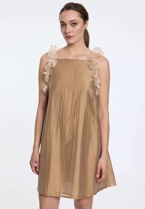 Femme portant une robe beige sans manches avec des bretelles transparentes à volants, debout devant un fond blanc uni.