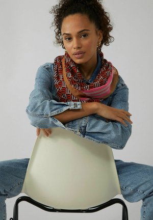 Multi-colored scarf featuring geometric patterns in red, pink, and brown, wrapped around the neck of a person in a denim jacket.