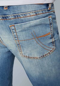 Jeans de denim de lavado claro con un bolsillo trasero con costuras naranjas y un patrón sutil. La tela tiene una textura ligeramente desgastada.