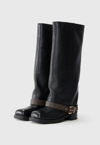 Zwarte leren kniehoge laarzen met een ronde neus, een platte zool en een decoratieve bruine riem met een gouden gesp rond de enkel.