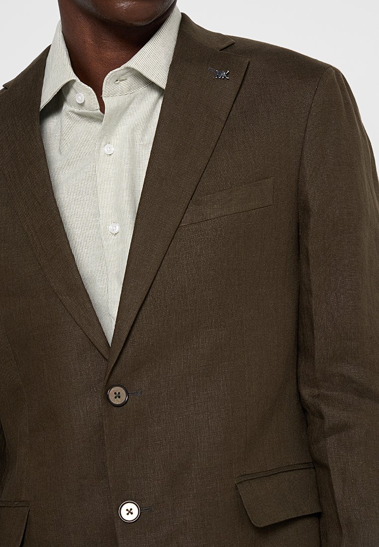 Brauner Stoff-Blazer mit strukturiertem Finish, ausgestattet mit einer einzelnen Brusttasche, zwei Knöpfen und einem hellgrünen gestreiften Hemd darunter.