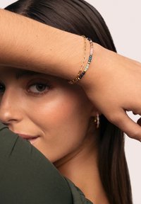 Guld kædearmbånd med flerfarvede perler i pink, blå, grøn og brun, båret på et håndled delvist skjult af en arm.